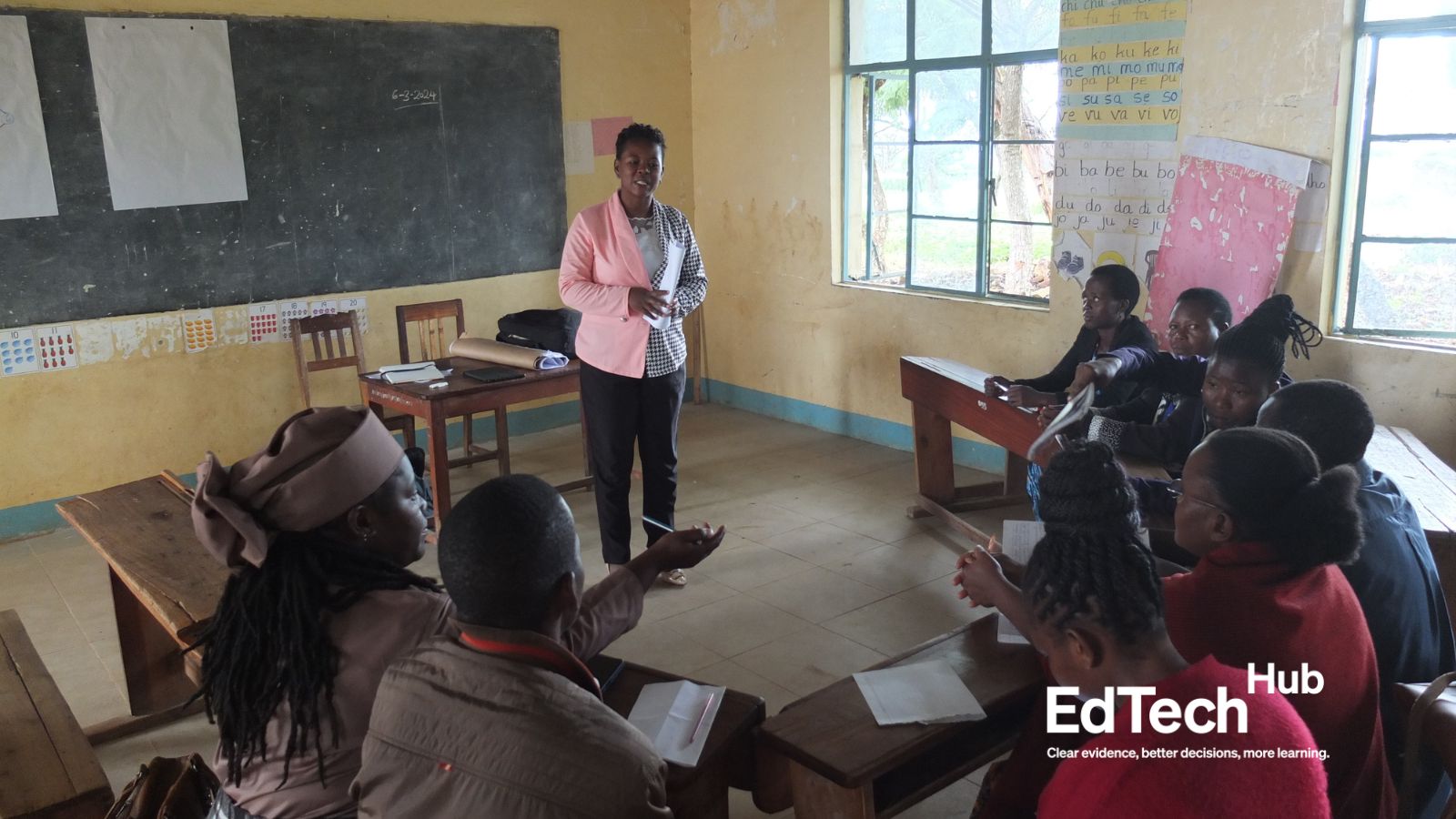 A researcher explains the next steps in a participatory activity to a group of teachers during a focus group discussion. Session is in a yellow schoolroom with chalkboard, desks, and manilla charts on the wall. Participants are seated at desks in a semi-circle around the researcher Photo credit: EdTech Hub Research team.