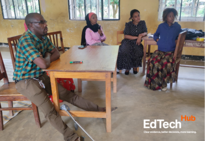 Teachers listen to a peer during a CoL session. Teachers are in a yellow classroom, seated in a semi-circle around a table. One teacher has space to stretch out his leg and a cane. Photo credit: Calvin Swai.