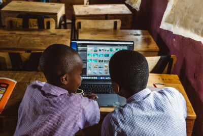 Two boys collaborate on a laptop in a classroom setting, sitting at a desk and focused on the screen.