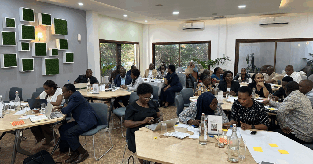 Photo from a workshop in Tanzania. Image shows room of stakeholders seated around 6 tables, working together using poster paper and post-it notes. 