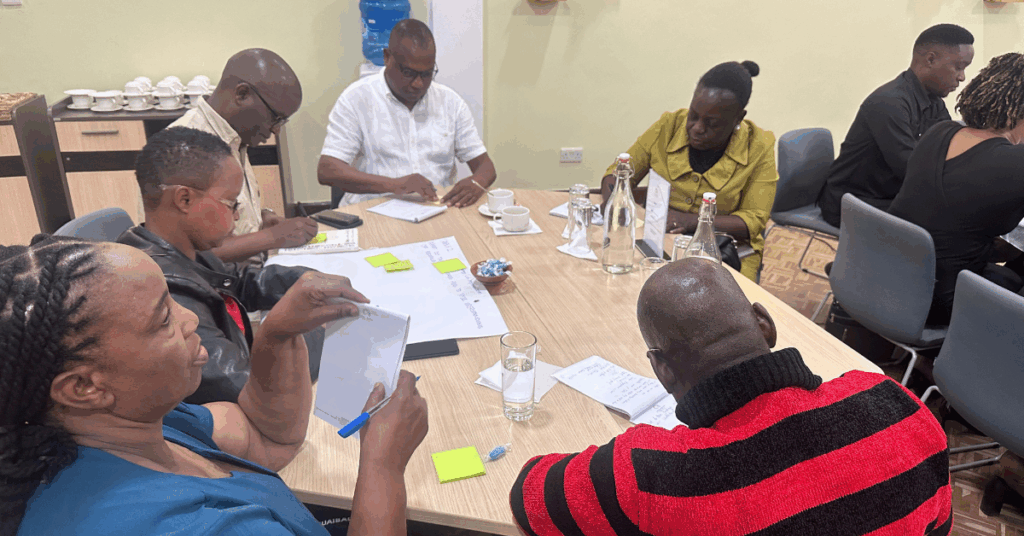 Photo from a workshop in Tanzania on Teacher Professional Development. Image shows group of 6 stakeholder participants seated around a rectangular table, writing on post-it notes and sticking them to a sheet of poster paper.